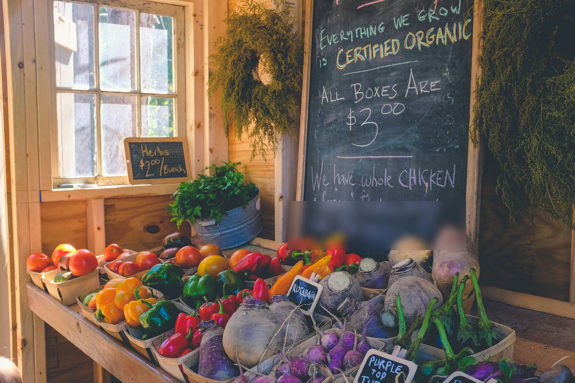 A farm-stand table stacked with baskets of peppers, tomatoes, and rutabagas beside a chalkboard advertising pasture-raised whole chicken